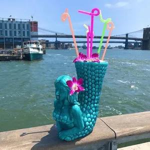 Mermaid-shaped tiki drink with colorful straws on a railing overlooking the East River and Brooklyn Bridge