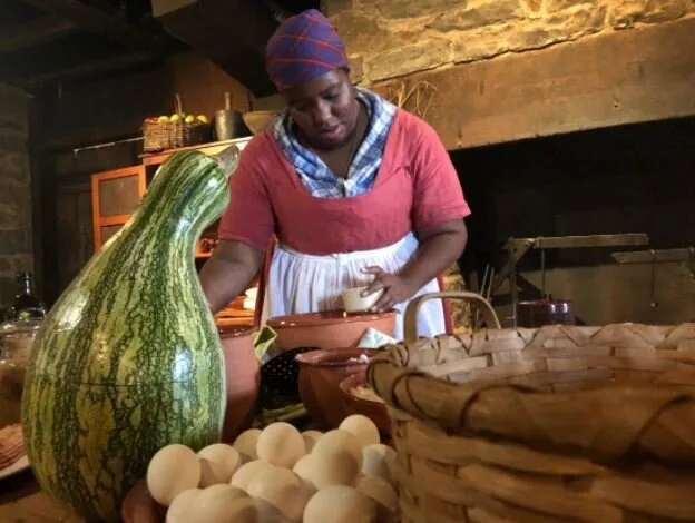 Costumed historical interpreter cooking at a hearth with gourds and baskets, from Not Your Momma's History