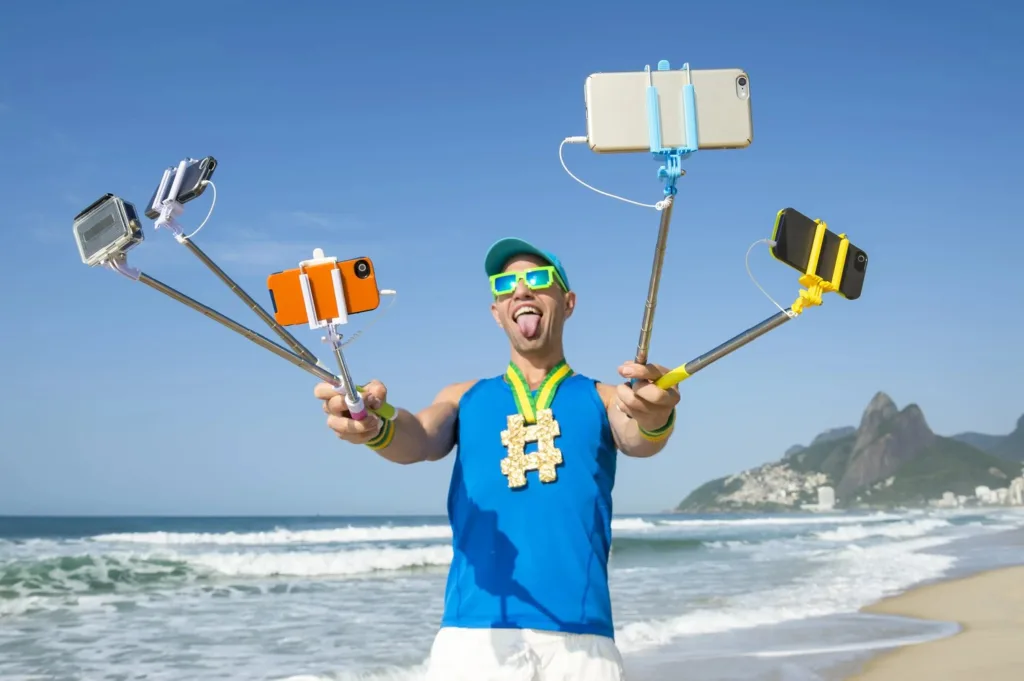 Man at the beach juggling several selfie sticks with phones and cameras, tongue out