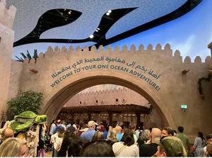 Crowd passing under a sandstone archway sign reading Welcome to Your One Ocean Adventure in Arabic and English