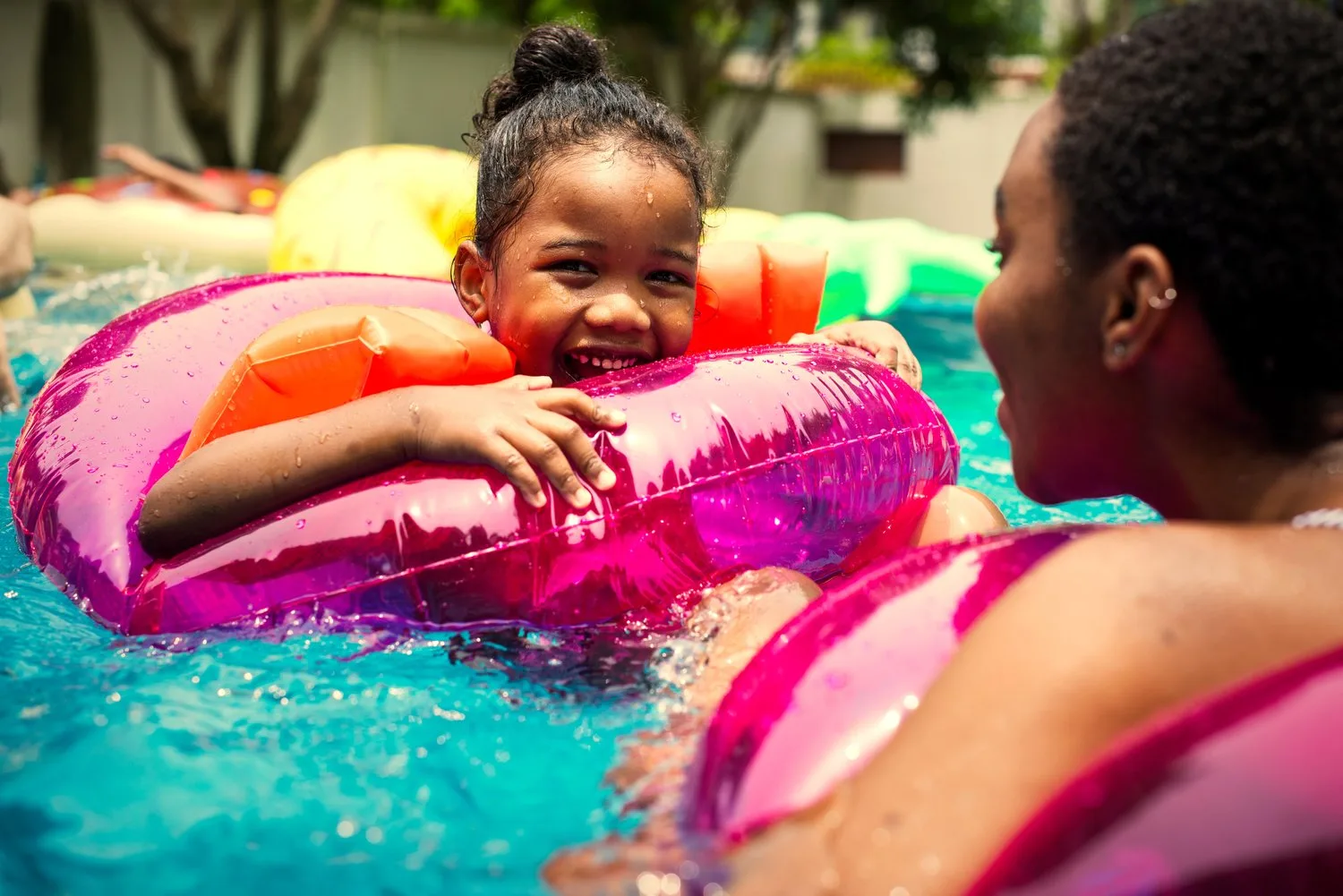 Smiling girl floating on a pink inner tube in a pool with a woman beside her