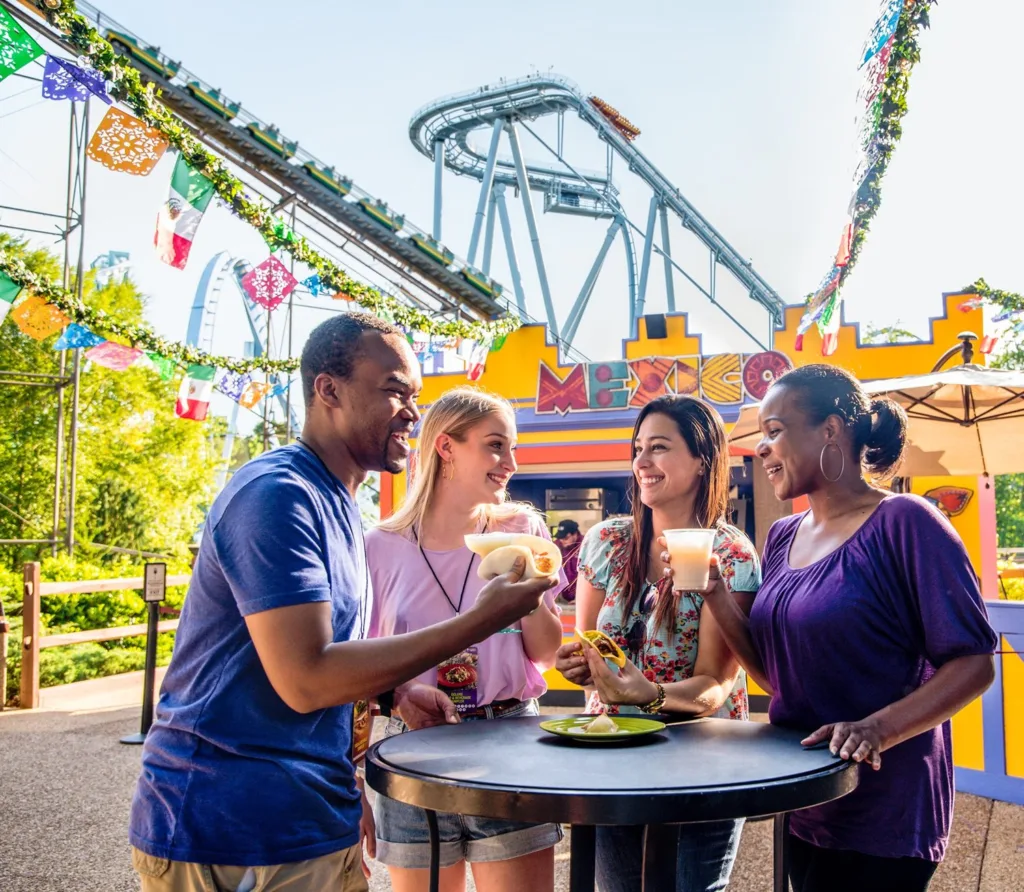 Four friends sharing tacos and drinks at a Mexico-themed food booth with a roller coaster behind at SeaWorld