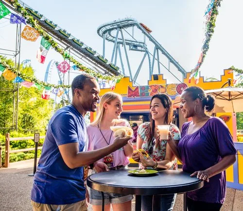 Friends sharing food and drinks at a high-top table at a festival booth near a roller coaster