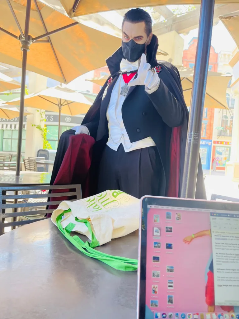 Costumed Dracula character posing by an outdoor table where a laptop is set up for remote work at a theme park