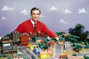 Mister Rogers' Neighborhood host in a red cardigan leaning over the show's miniature neighborhood model