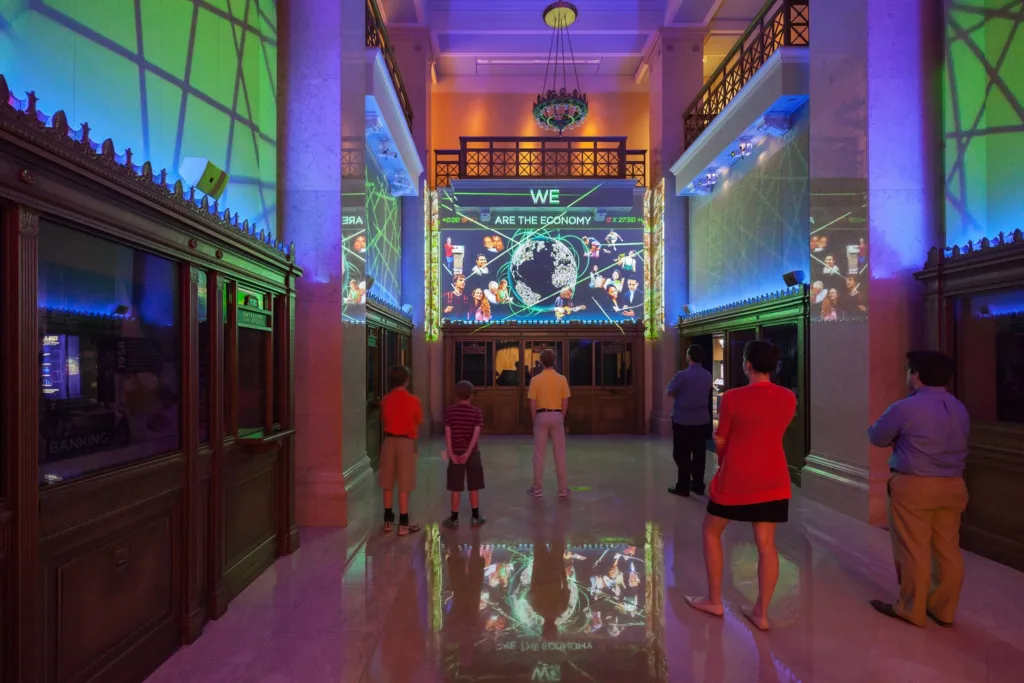Visitors in a grand bank-style hall with We Are the Economy projections on the walls