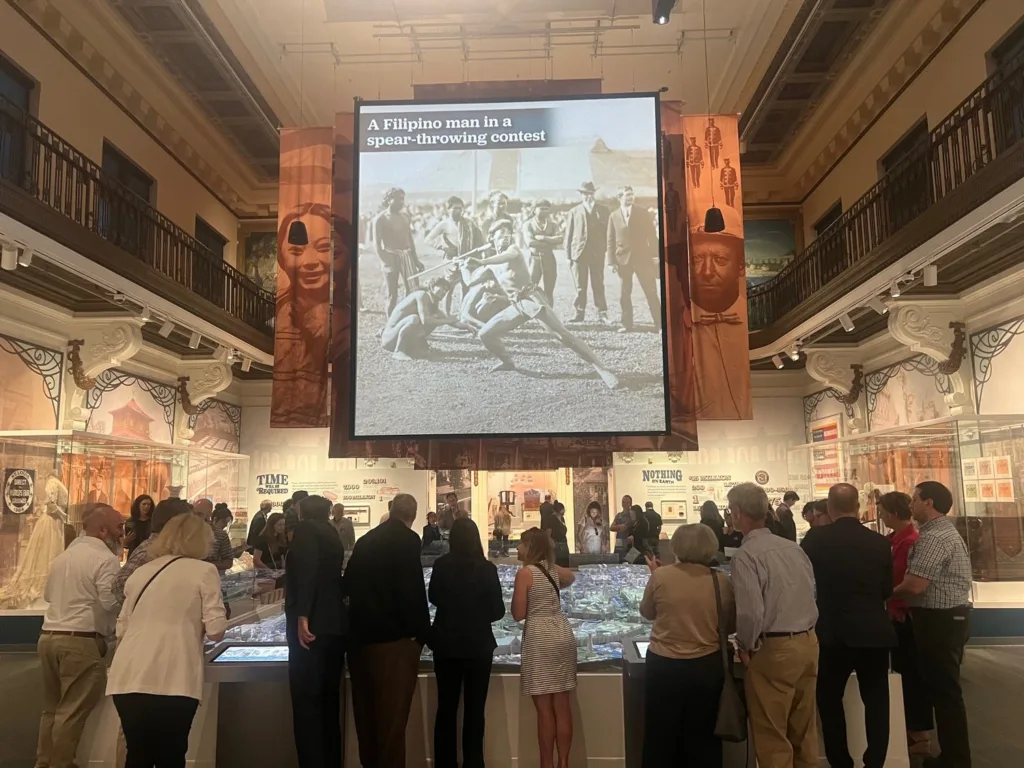 Visitors gathered around the 1904 World's Fair model at the Missouri History Museum beneath a projected historic photo