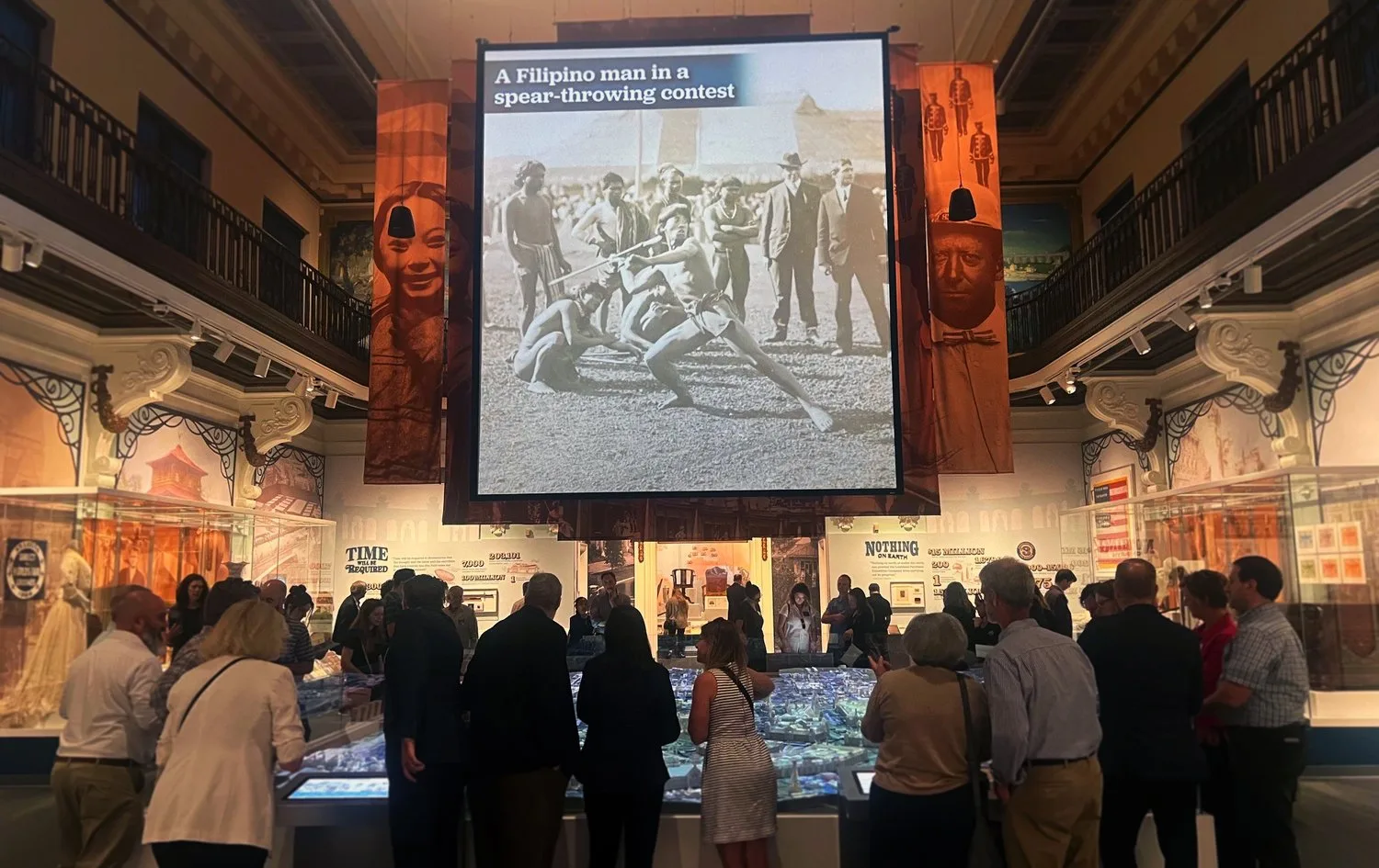 Visitors gathered around the 1904 World's Fair model at the Missouri History Museum under a projected archival photo