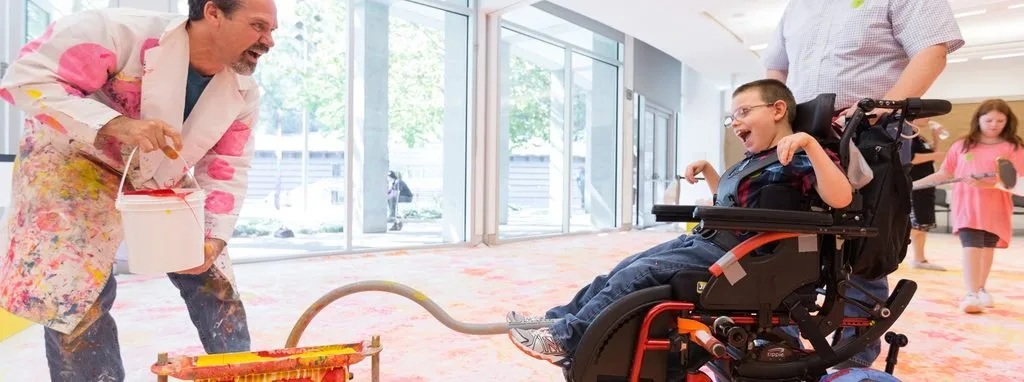 Artist in a paint-splattered coat helping a child in a wheelchair create paint art on the floor