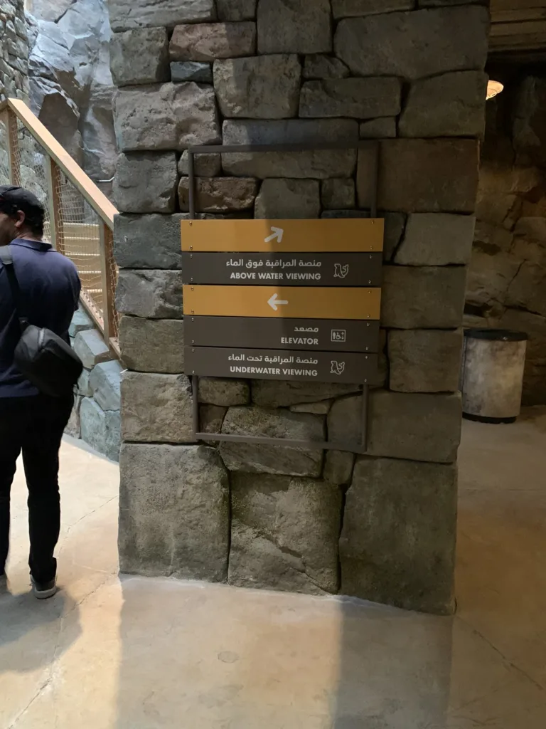 Bilingual Arabic and English directional signs for above water viewing, elevator and underwater viewing on a rockwork pillar at SeaWorld Abu Dhabi