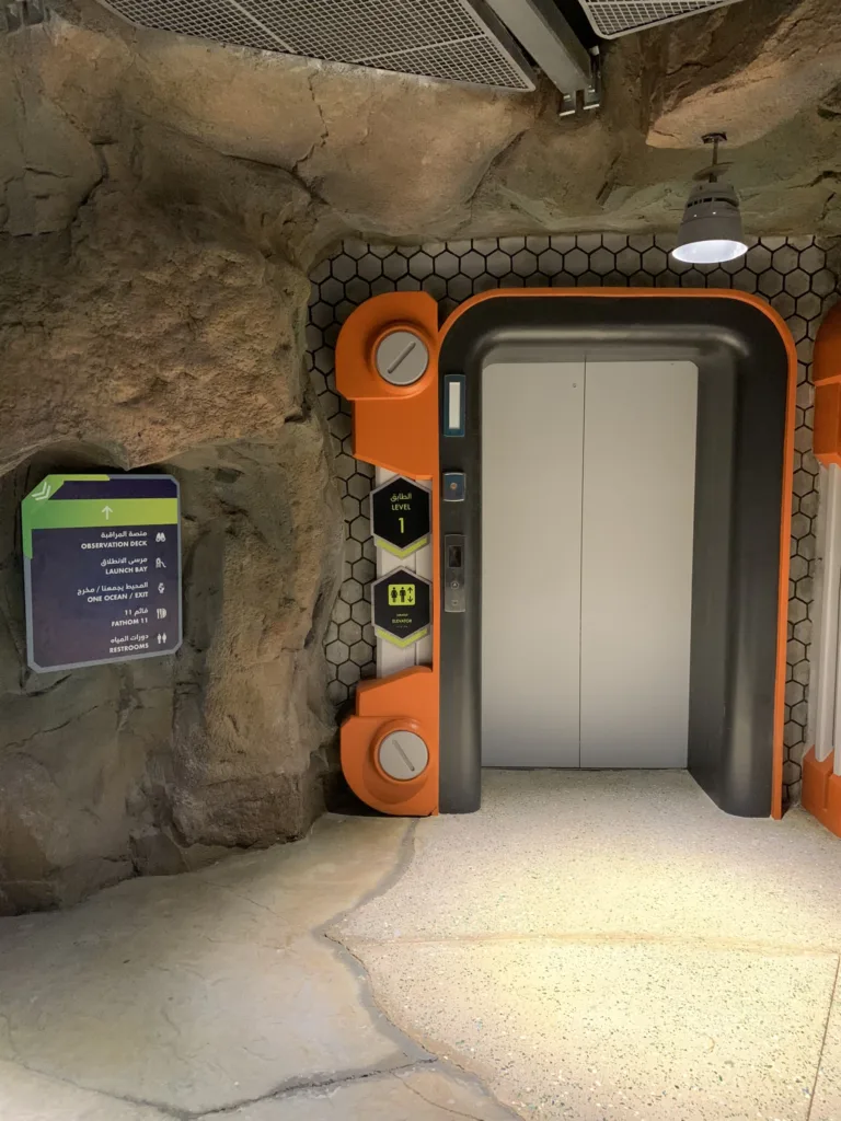 Elevator in a rockwork wall with an orange sci-fi frame and a wayfinding sign in the Endless Ocean realm at SeaWorld Abu Dhabi