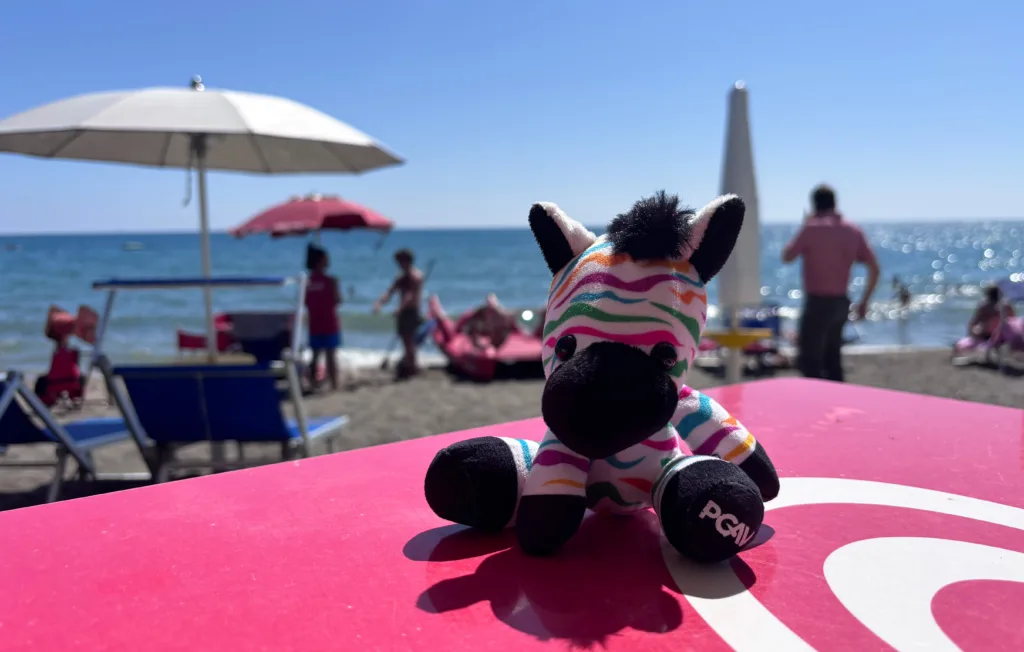 Spot the PGAV zebra plush on a pink table at a sunny beach with umbrellas and swimmers