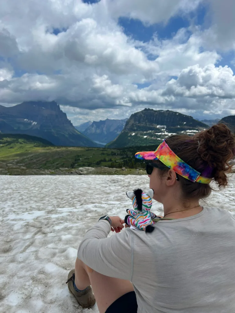 Hiker with Spot the PGAV zebra plush on her shoulder overlooking snowfields and peaks at Glacier National Park