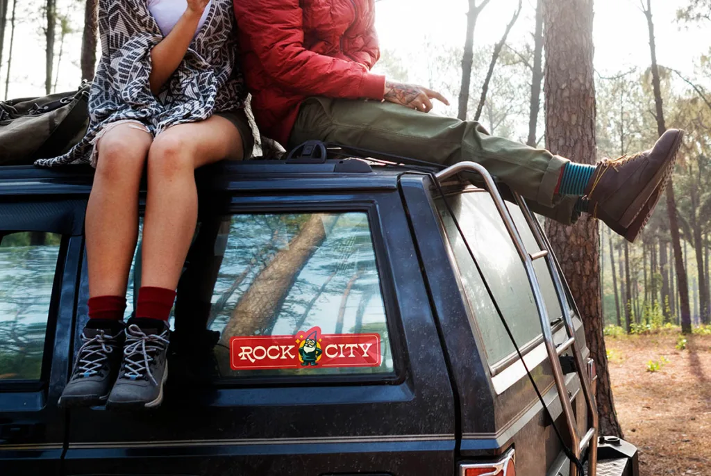 Rock City gnome logo sticker on the back window of an SUV where two travelers sit on the roof in a forest