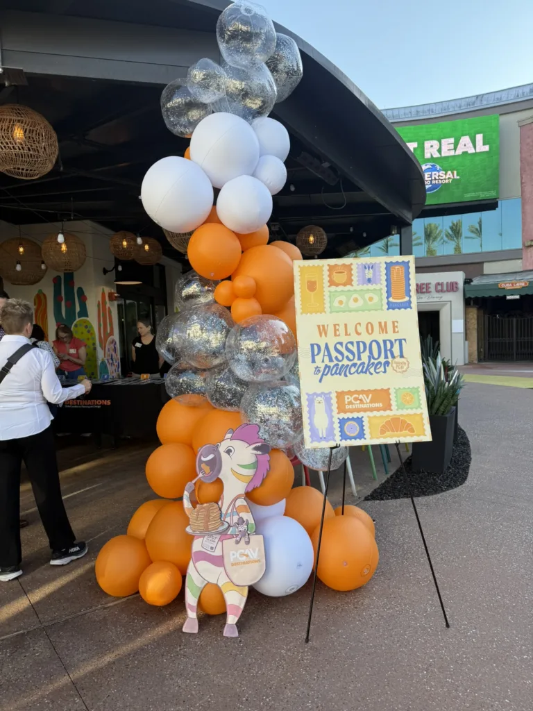 Orange, white and clear balloon column beside a Passport and Pancakes welcome sign at the PGAV pancake breakfast
