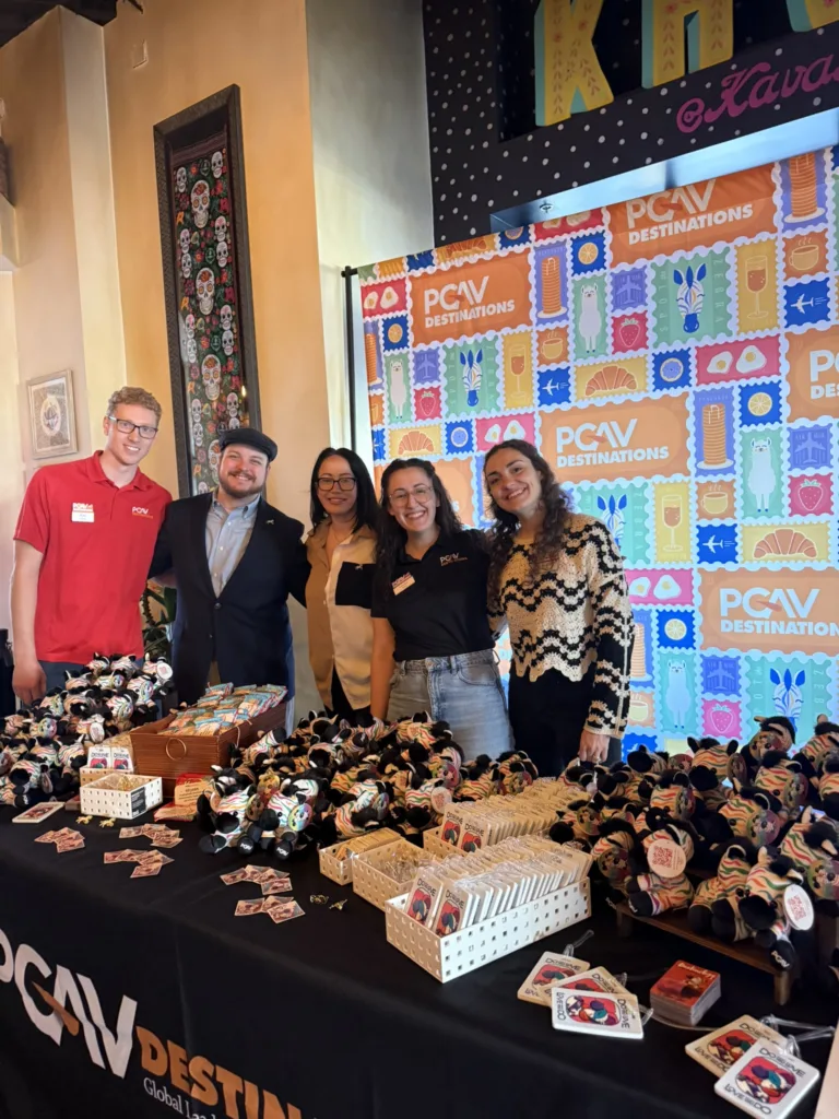 PGAV team members at a swag table of plush toys and pins in front of a colorful stamp-pattern backdrop