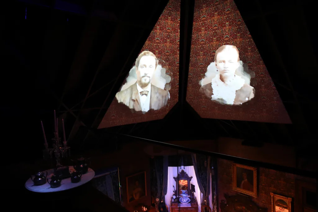 Historic portraits of a couple projected on angled screens over a darkened Victorian room in the Schifferdecker and Zelleken homes