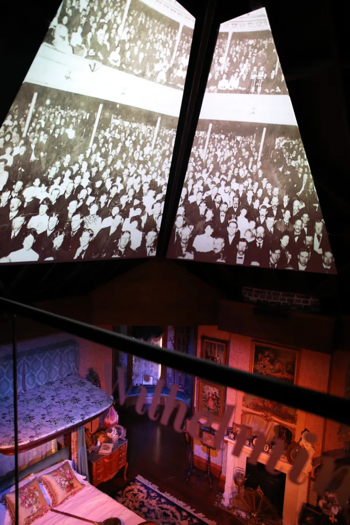 Projected historic crowd photos on angled screens above a lit Victorian bedroom seen from the observation deck