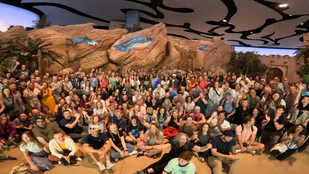 Large group of PGAV team members waving in a group photo inside a themed attraction with rockwork and shark murals